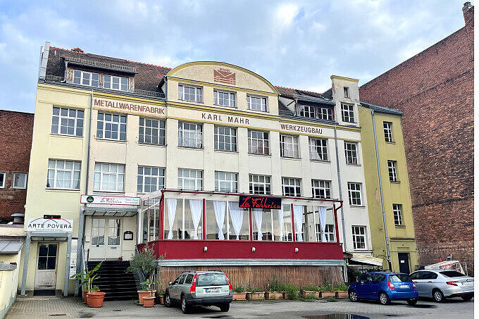 Aufnahme eines großen Hauses in Gostenhof mit der Aufschrift 'Karl Mahr Metallwarenfabrik und Werkzeugbau', im Erdgeschoss ein Restaurant mit Wintergarten, vor dem Haus parkende Autos